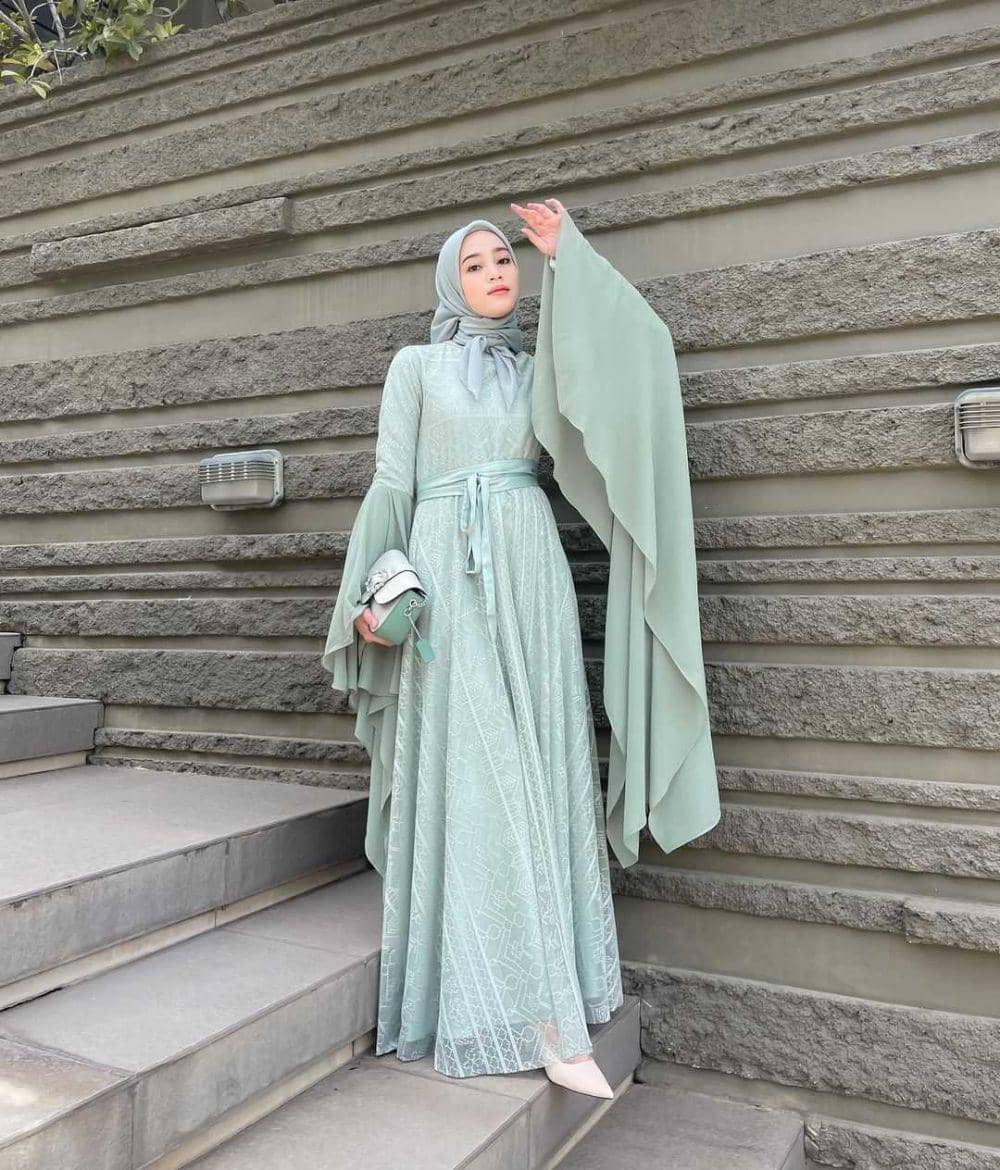 Seorang wanita berhijab mengenakan gaun panjang berwarna hijau mint dengan lengan lebar, berdiri di tangga dekat dinding batu abu-abu.