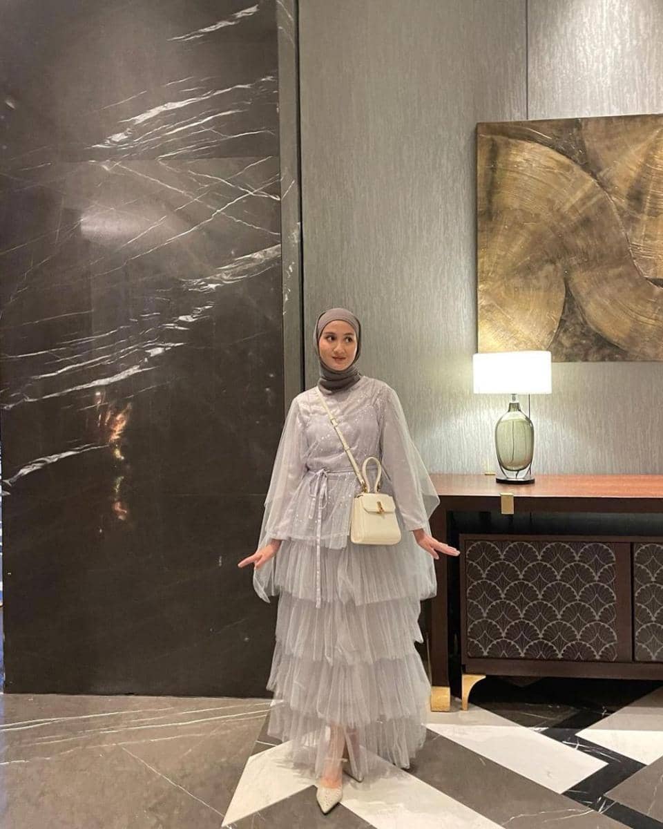 Seorang wanita berhijab mengenakan gaun tulle berlapis warna abu-abu muda dengan tas putih, berdiri di lobi hotel bergaya modern.