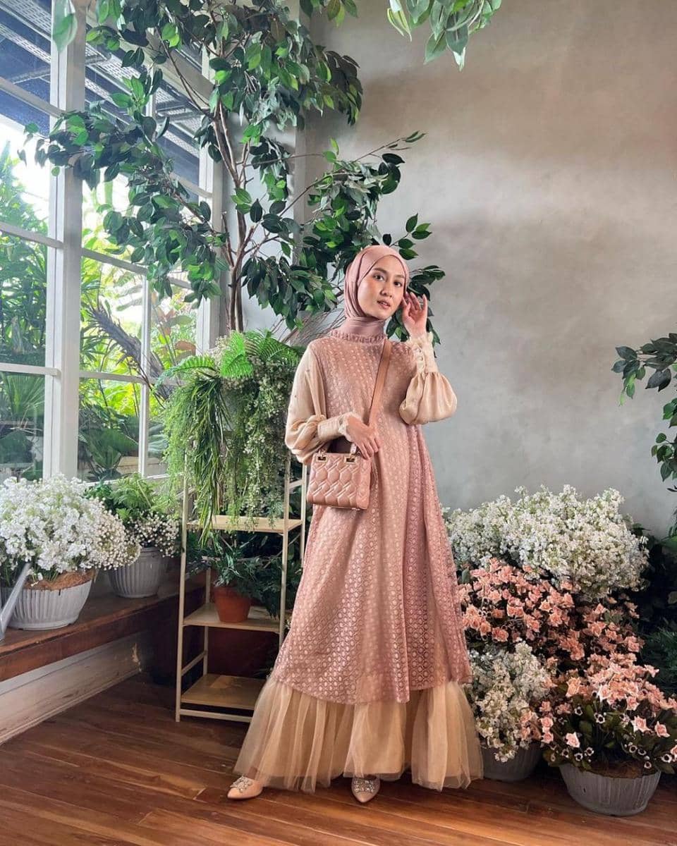 Seorang wanita berhijab mengenakan gaun panjang berwarna pastel dan tas kecil berwarna senada, berpose di ruangan penuh tanaman hias dan bunga.
