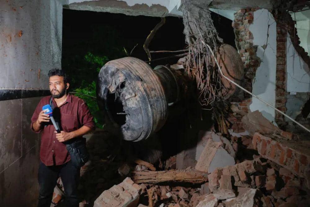 Seorang jurnalis berdiri di sebelah puing-puing roda pendaratan pesawat Air India AI171 (Dok. Reuters)