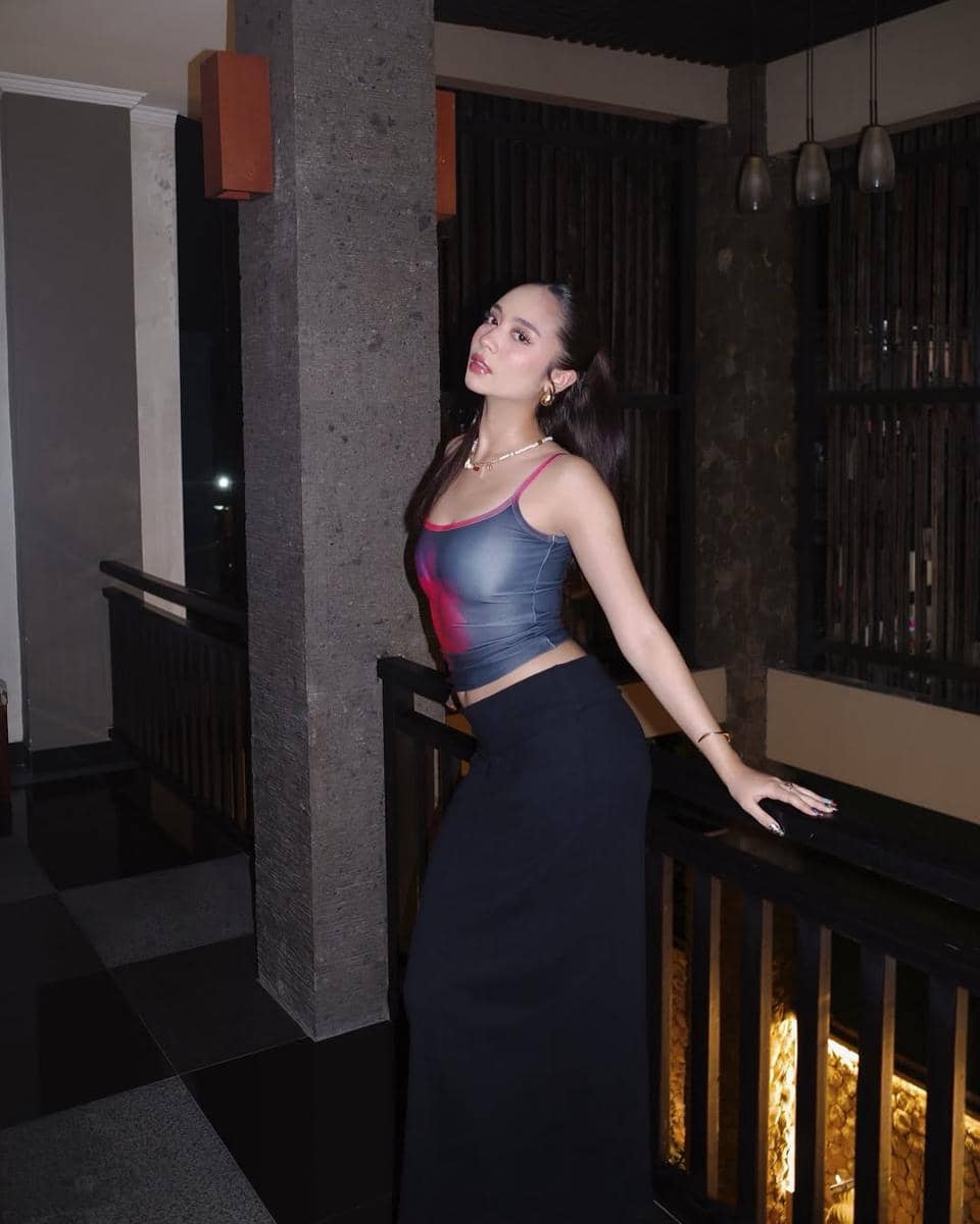 Seorang perempuan berpose di dalam ruangan dengan pencahayaan redup, mengenakan tank top abu-abu dan rok panjang hitam.
