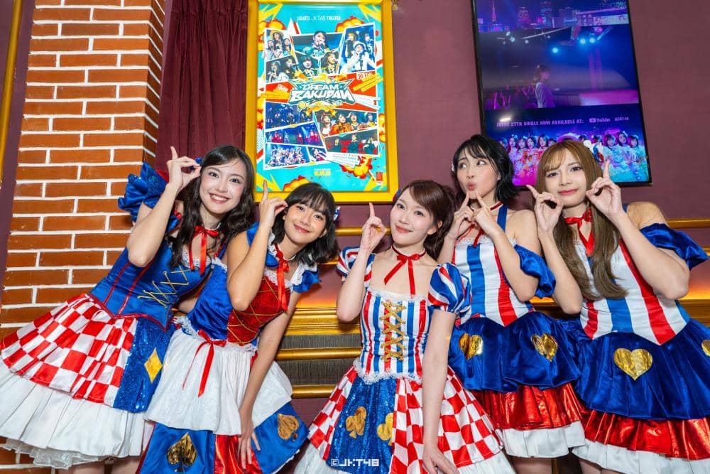 Lima anggota grup idola JKT48 berpose ceria mengenakan kostum panggung berwarna merah, putih, dan biru dengan latar dinding bata dan poster acara.