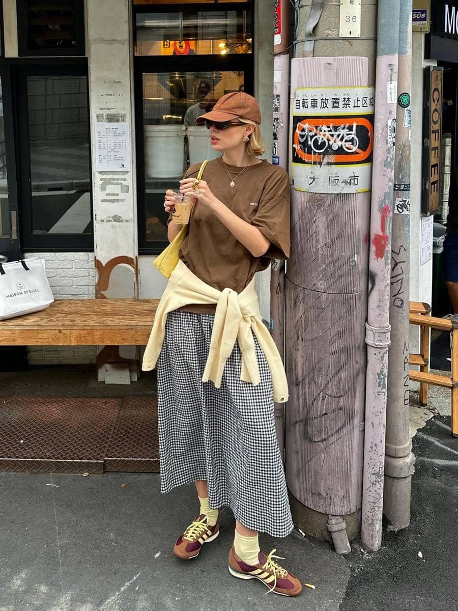 Seorang wanita bergaya kasual mengenakan kaus cokelat, rok kotak-kotak, topi, dan sepatu sneakers sambil memegang minuman di jalan kota.