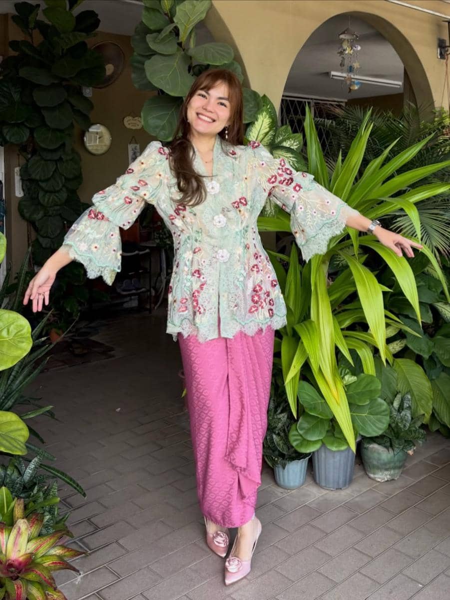 Seorang wanita tersenyum mengenakan kebaya hijau muda bermotif bunga dan kain batik pink, berpose di depan tanaman hias di halaman rumah.