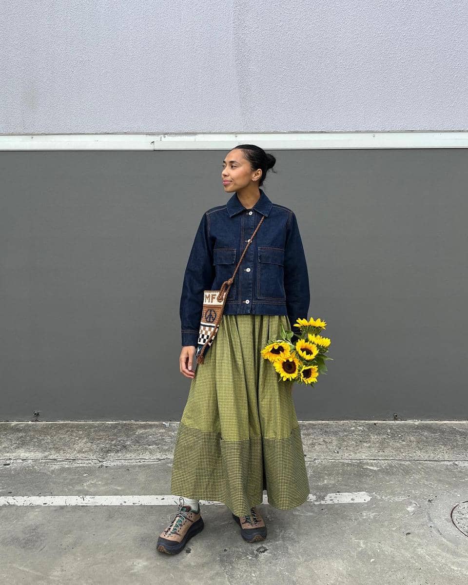 Seorang wanita berdiri di luar ruangan mengenakan jaket denim biru tua, rok panjang hijau bermotif kotak, dan memegang buket bunga matahari.