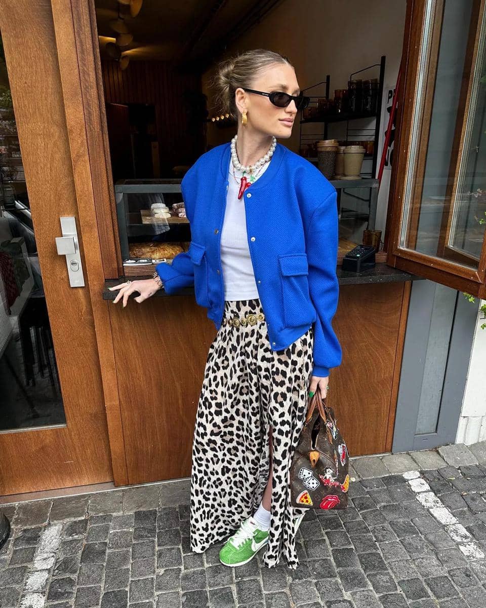 Seorang wanita bergaya mengenakan jaket biru cerah, rok motif leopard, sepatu hijau, dan kacamata hitam berdiri di depan kafe kayu.