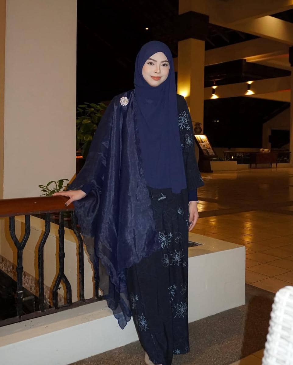 Seorang wanita mengenakan hijab dan busana berwarna biru tua dengan motif bunga berdiri di area dalam gedung yang diterangi lampu malam.