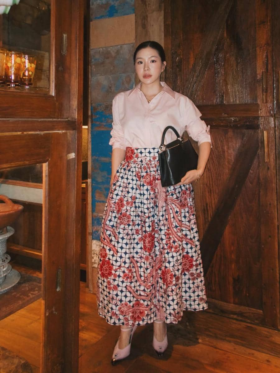 Seorang wanita mengenakan kemeja berwarna merah muda dan rok panjang bermotif batik merah biru sambil memegang tas hitam di ruangan kayu.