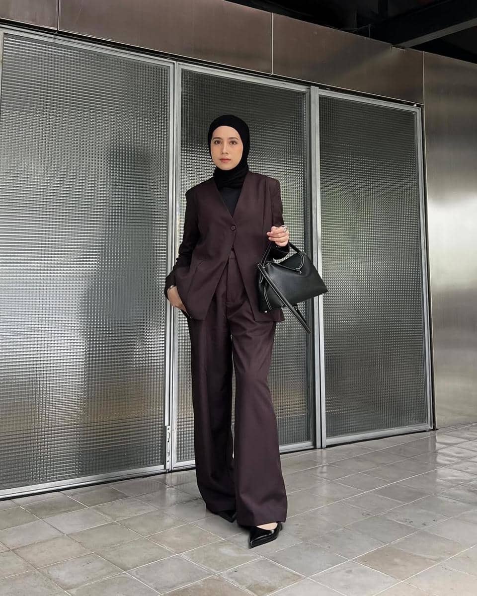 Seorang wanita berhijab mengenakan setelan jas berwarna cokelat tua dengan celana panjang lebar dan membawa tas hitam di depan dinding logam modern.