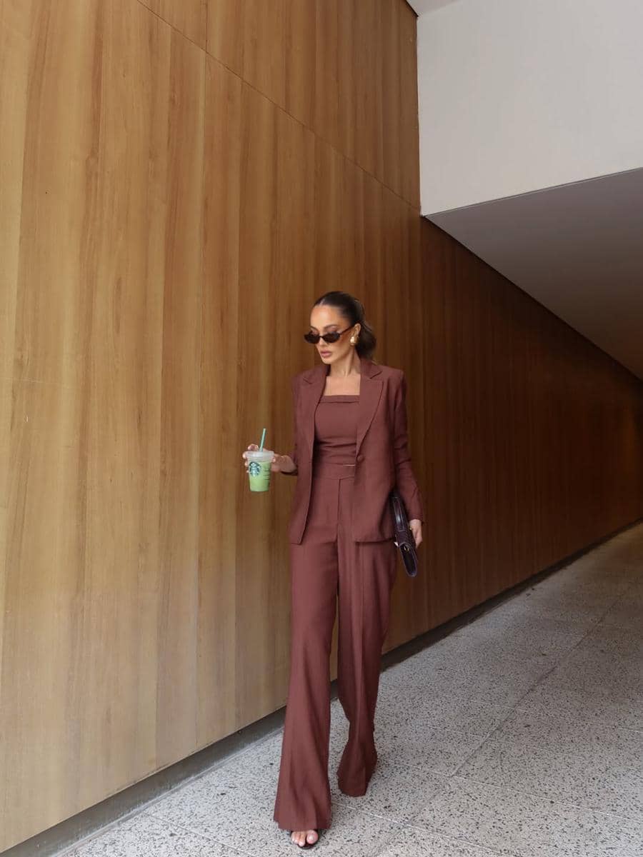 Seorang wanita mengenakan setelan blazer dan celana panjang berwarna cokelat berjalan di lorong dengan dinding kayu sambil memegang minuman hijau.