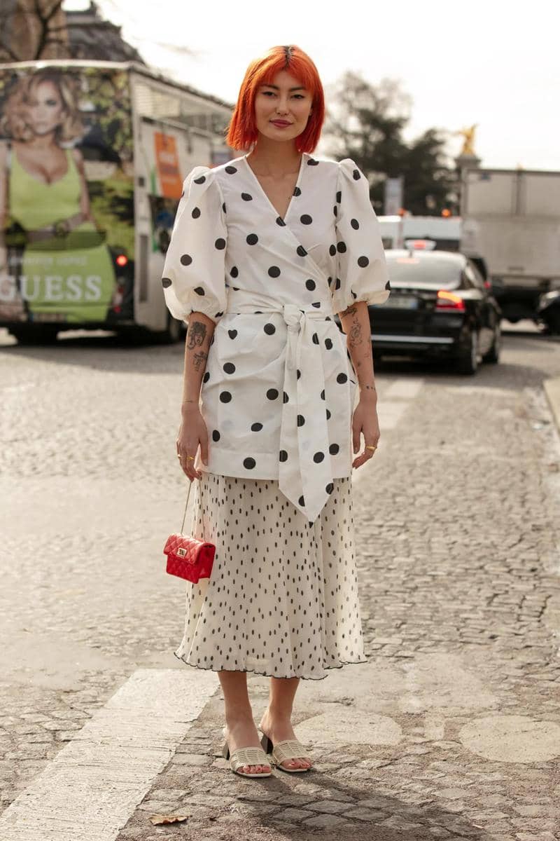 Seorang wanita berambut oranye mengenakan blus putih bermotif polkadot besar dan rok midi bermotif titik kecil di jalan kota.