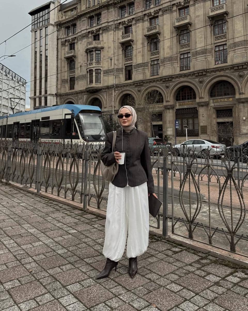 Seorang wanita berhijab bergaya modis mengenakan atasan gelap dan celana putih longgar berdiri di trotoar kota dengan latar bangunan klasik dan trem.