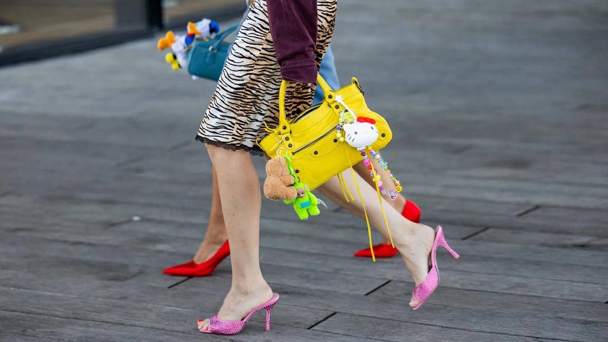 Dua wanita berjalan mengenakan sepatu hak tinggi berwarna cerah, membawa tas tangan kuning dan biru dengan gantungan boneka lucu.