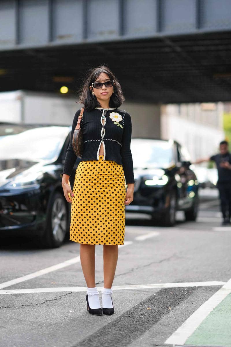 Seorang wanita bergaya mengenakan rok kuning bermotif polkadot hitam dan atasan rajut hitam dengan hiasan bunga putih di jalan kota.