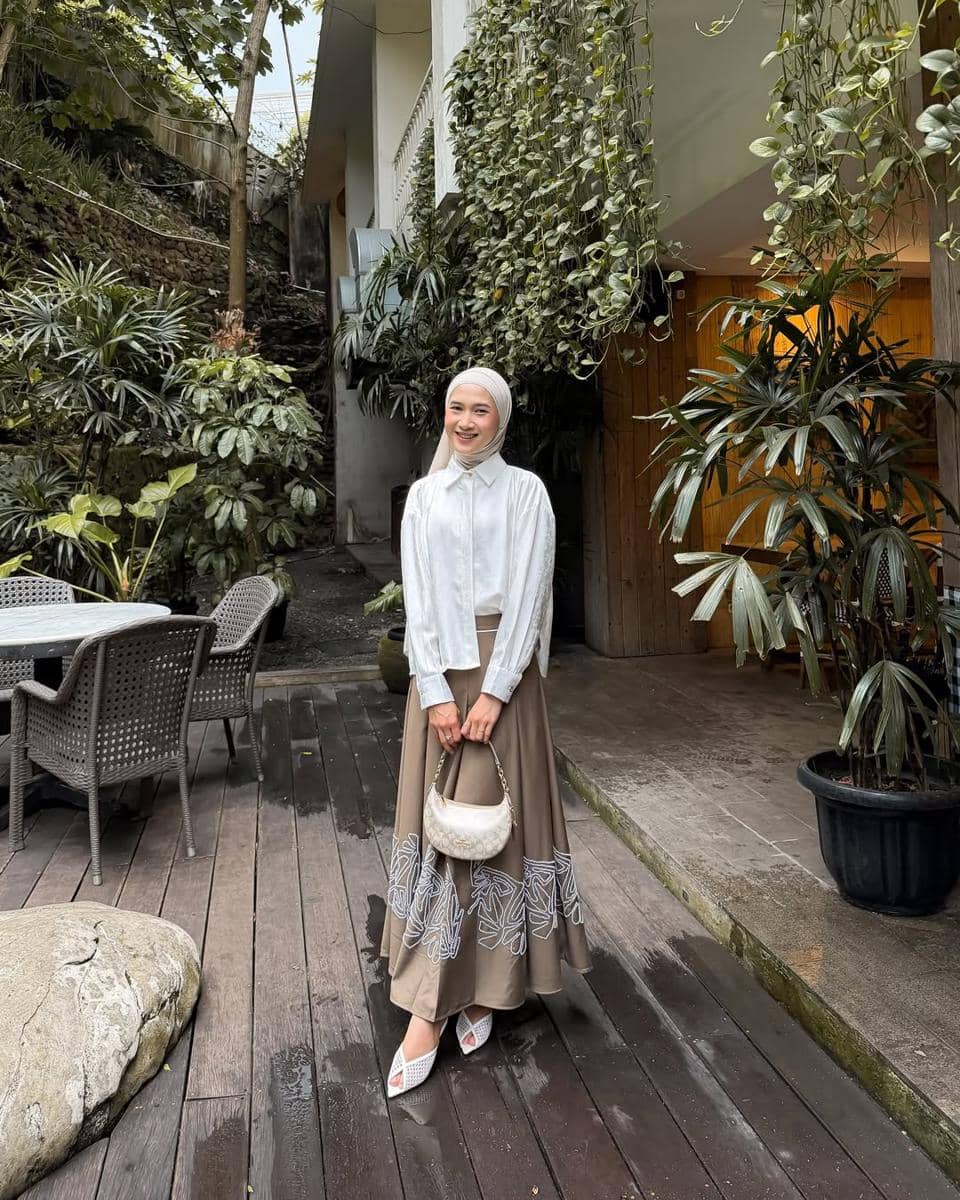 Seorang wanita berhijab mengenakan kemeja putih dan rok panjang cokelat bermotif berdiri di teras kayu dengan latar tanaman hijau.