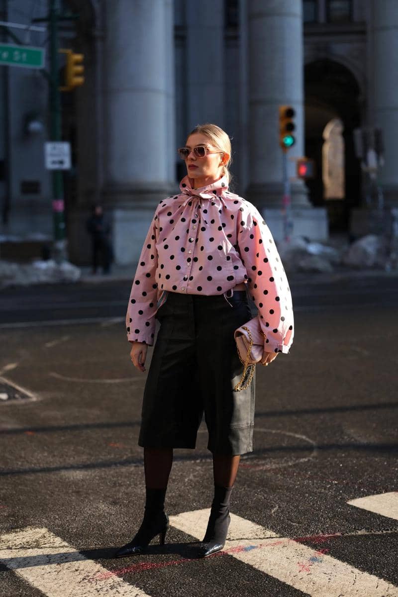 Seorang wanita bergaya mengenakan blus merah muda bermotif polkadot hitam, celana kulot hitam, dan kacamata di jalan kota.