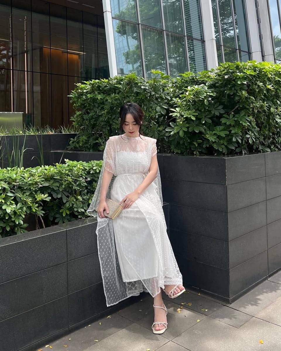 Seorang wanita mengenakan gaun putih elegan duduk di tepi taman dengan latar gedung kaca modern dan tanaman hijau di sekitarnya.