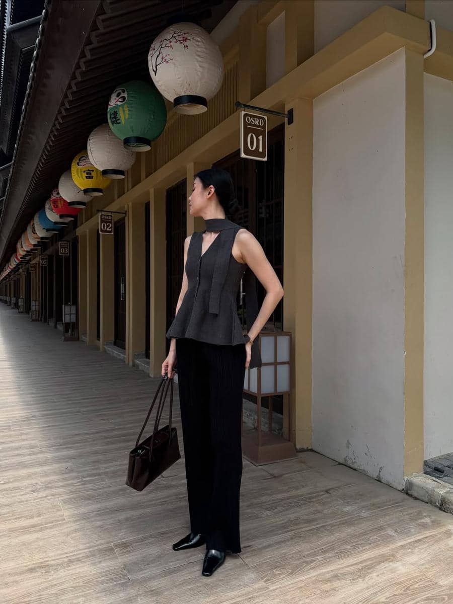 Seorang wanita bergaya elegan mengenakan atasan tanpa lengan dan celana panjang hitam sambil membawa tas di lorong dengan lentera Jepang.