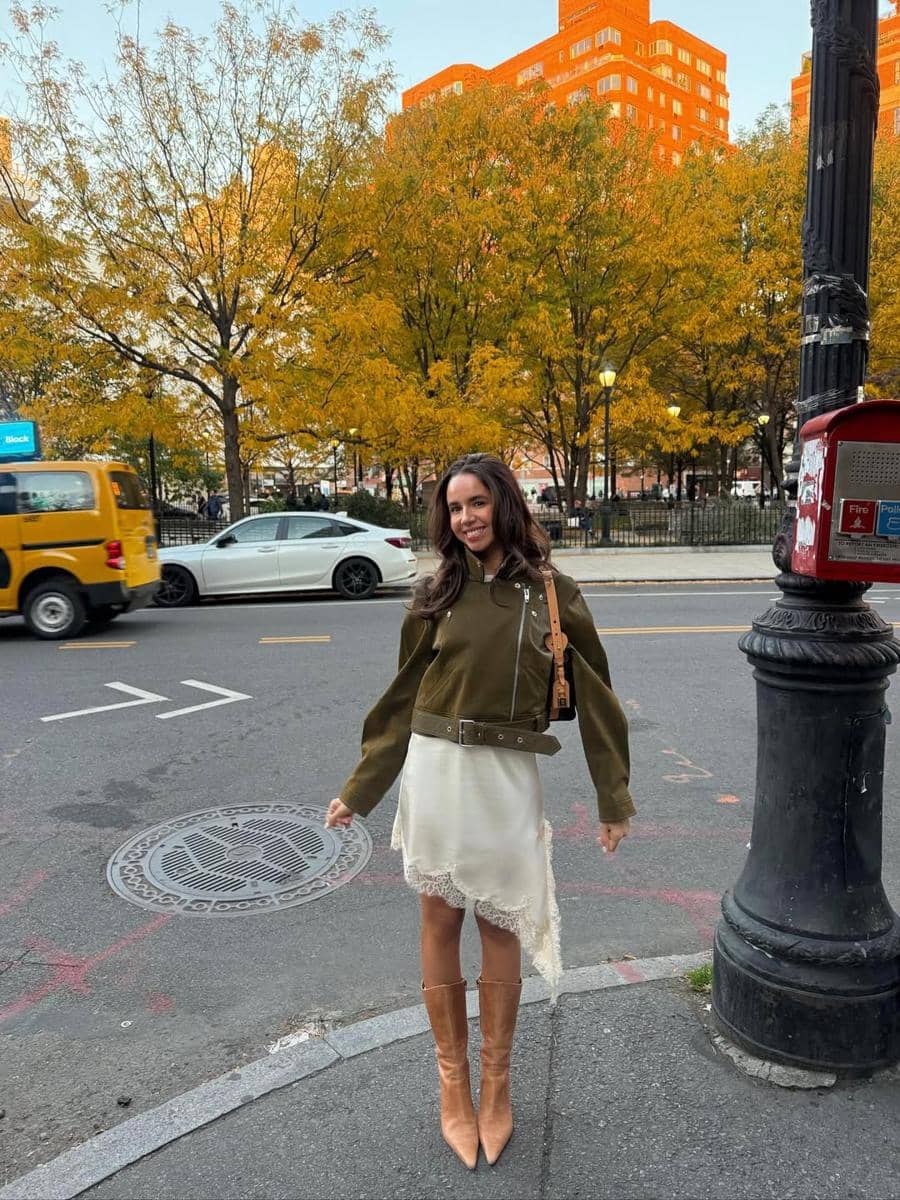 Seorang wanita berdiri di tepi jalan kota dengan latar pepohonan berdaun kuning musim gugur dan gedung bata oranye di kejauhan.