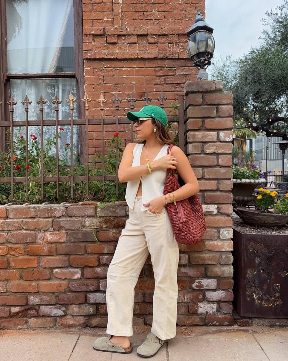 Seorang wanita mengenakan topi hijau, rompi putih, dan celana krem sambil membawa tas rajut merah di depan dinding bata tua.