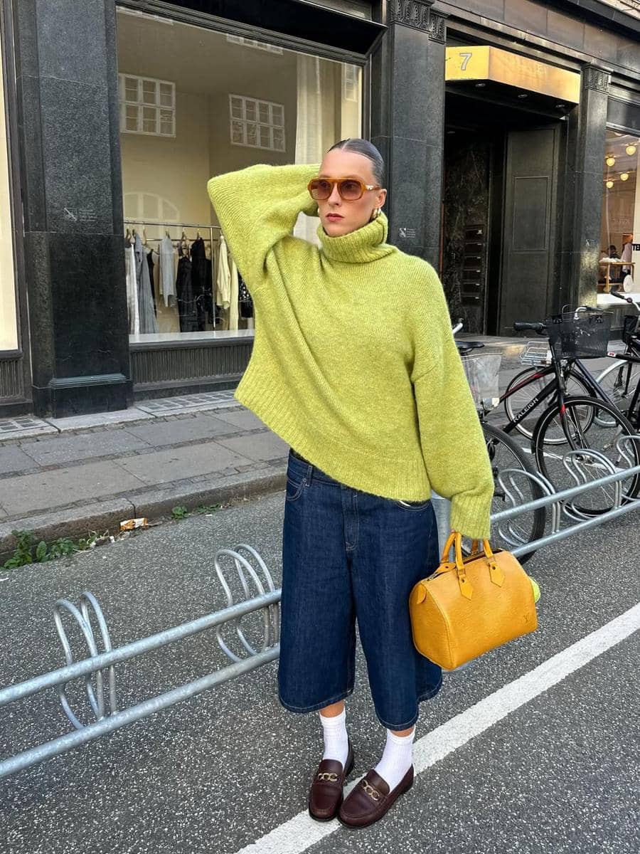 Seorang wanita mengenakan sweater hijau terang, celana denim panjang selutut, dan kacamata cokelat besar sambil memegang tas kuning di trotoar kota.