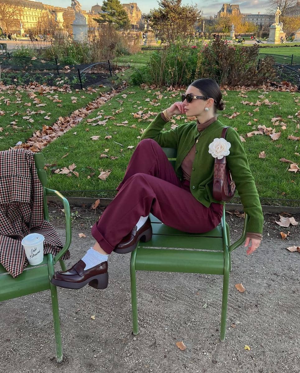 Seorang wanita bergaya duduk santai di kursi taman dengan mengenakan kardigan hijau, celana marun, dan sepatu hak cokelat.