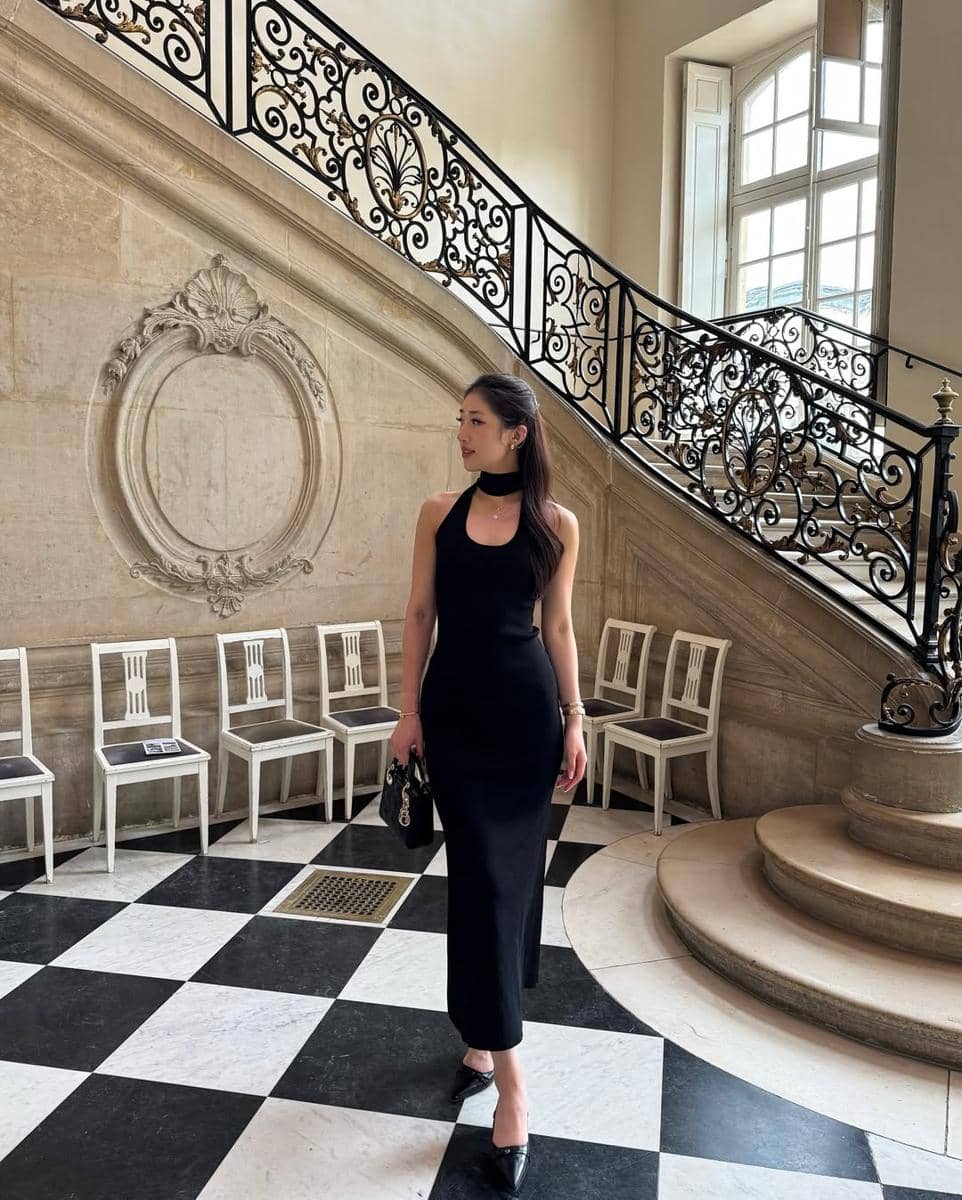 Seorang wanita mengenakan gaun hitam elegan berdiri di ruangan bergaya klasik dengan tangga besar dan lantai kotak hitam putih.