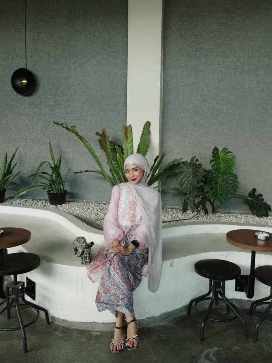 Seorang wanita berhijab duduk di kafe dengan latar dinding abu-abu dan tanaman hias, mengenakan pakaian bernuansa pink dan ungu.