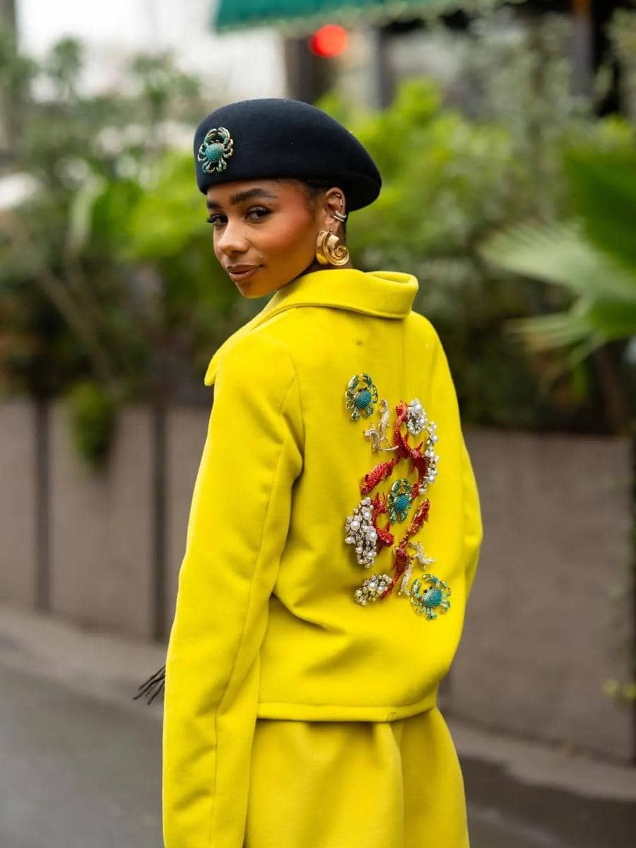 Seorang wanita mengenakan mantel kuning cerah dengan hiasan permata di bagian belakang dan topi baret hitam berhias bros biru.