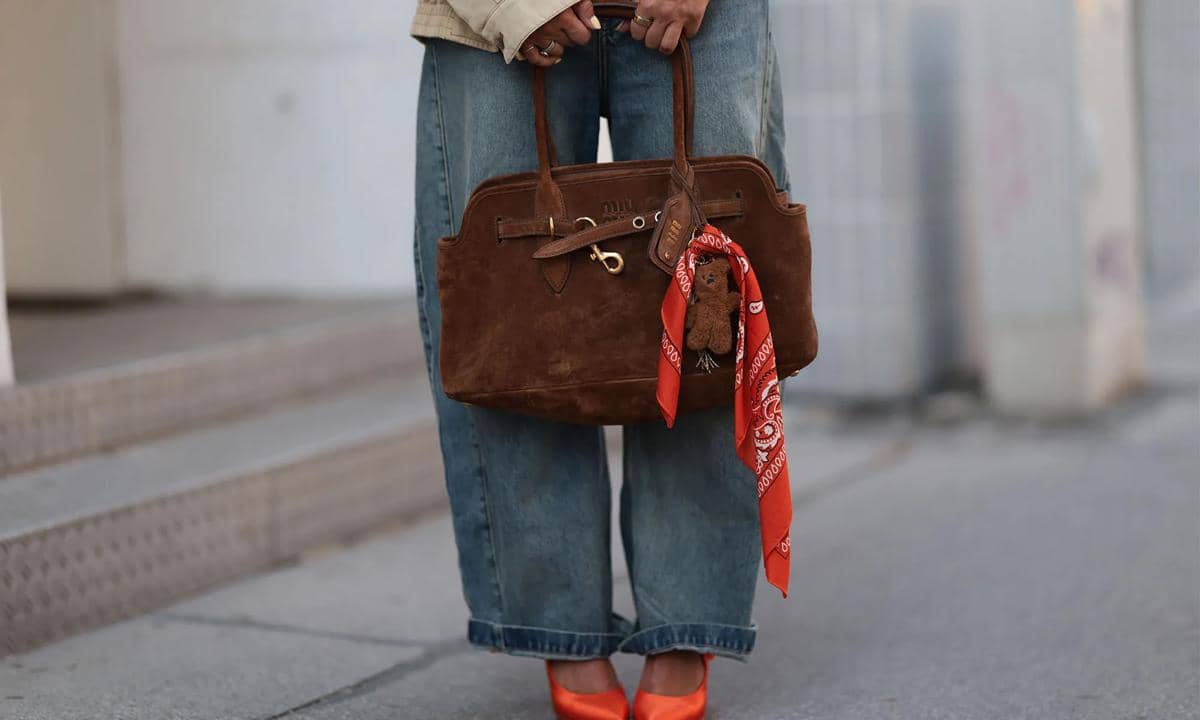 Tas cokelat berbahan suede dengan gantungan boneka kecil dan syal merah, dipadukan dengan celana jeans longgar dan sepatu hak tinggi oranye.