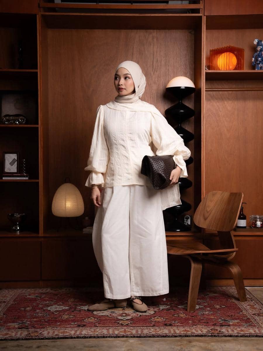 Seorang wanita berhijab mengenakan atasan dan celana berwarna krem sambil memegang tas tangan di ruangan bergaya retro dengan furnitur kayu.