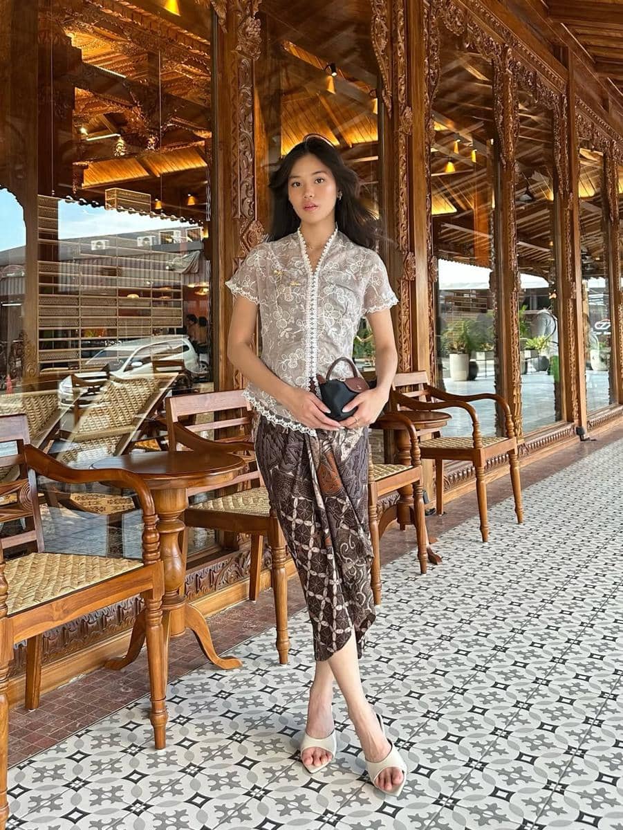 Seorang wanita mengenakan kebaya putih dan kain batik berdiri di depan bangunan kayu berukir dengan kursi rotan di teras.