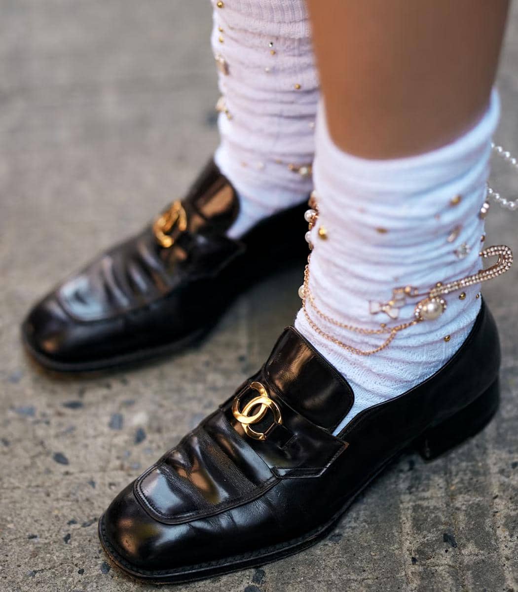 Sepasang sepatu loafer hitam dengan detail logam emas dipadukan dengan kaus kaki putih berhias mutiara dan rantai emas.