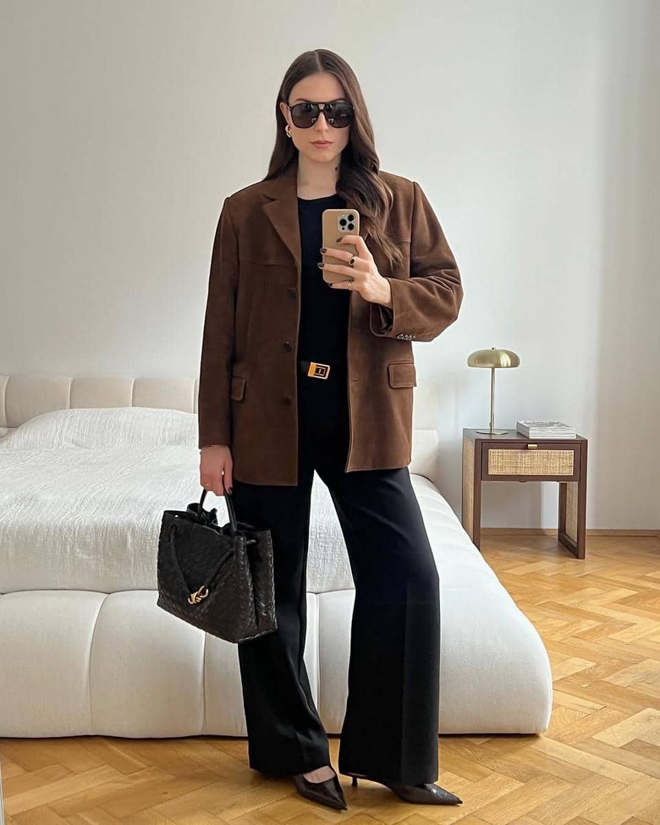 Seorang wanita bergaya elegan mengenakan blazer cokelat, celana hitam, dan kacamata hitam sambil berpose di kamar minimalis.
