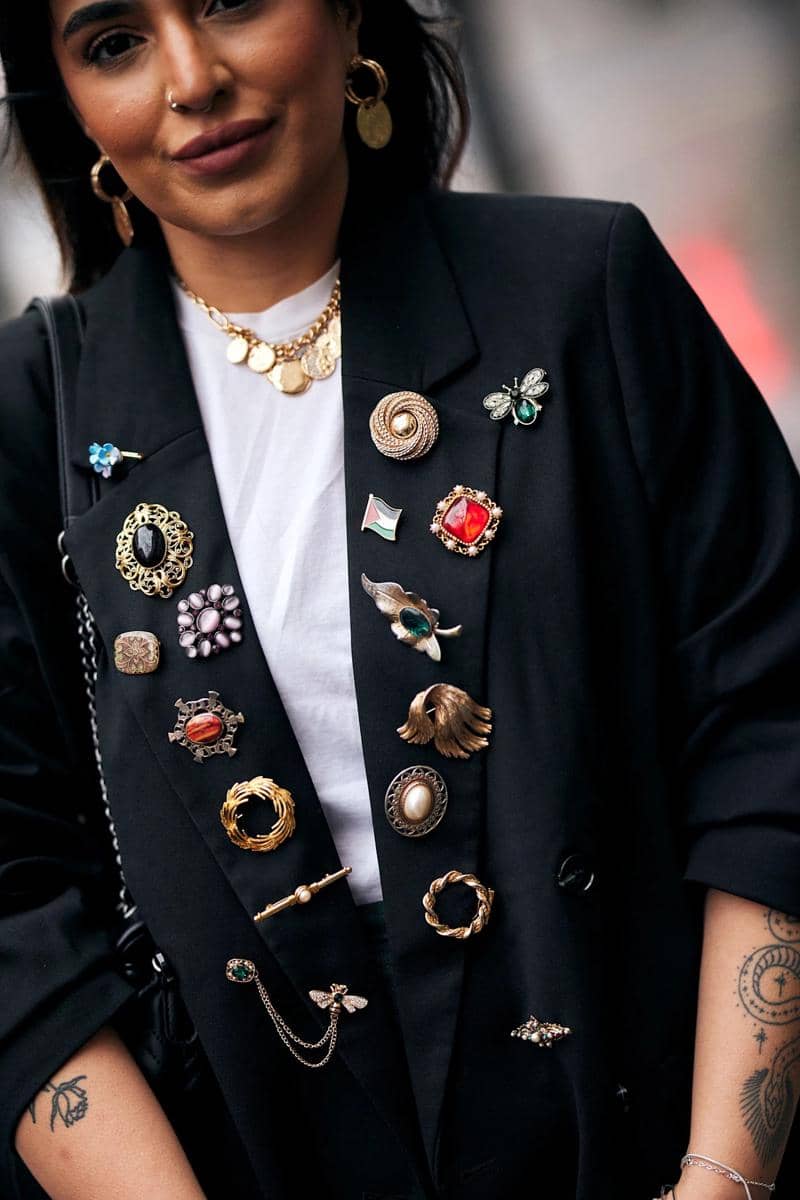 Seorang wanita mengenakan blazer hitam berhias berbagai bros vintage berwarna emas dan perak, dipadukan dengan kaus putih dan perhiasan emas.
