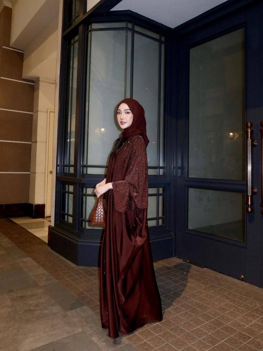 Seorang wanita berhijab mengenakan gaun panjang berwarna marun dengan tas cokelat berdiri di depan pintu kaca besar pada malam hari.