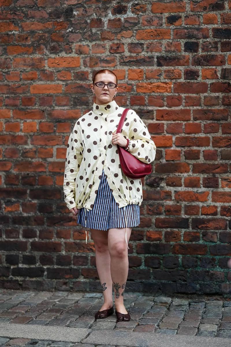 Seorang wanita bergaya mengenakan blus kuning bermotif polkadot besar, rok mini bergaris biru putih, dan tas bahu merah di depan dinding bata.