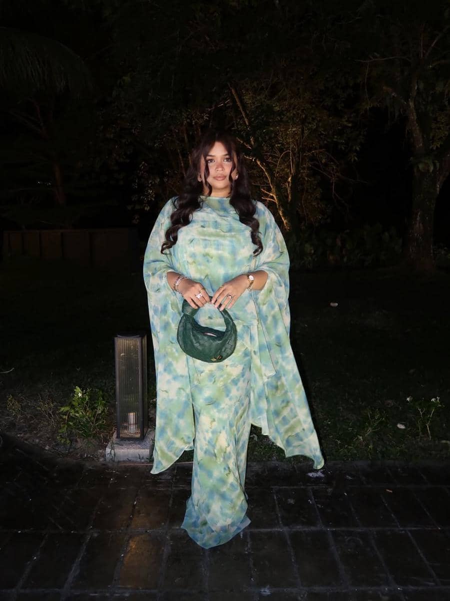 Seorang wanita mengenakan saree bermotif hijau dan biru dengan tas tangan hijau, berdiri di area luar ruangan pada malam hari.