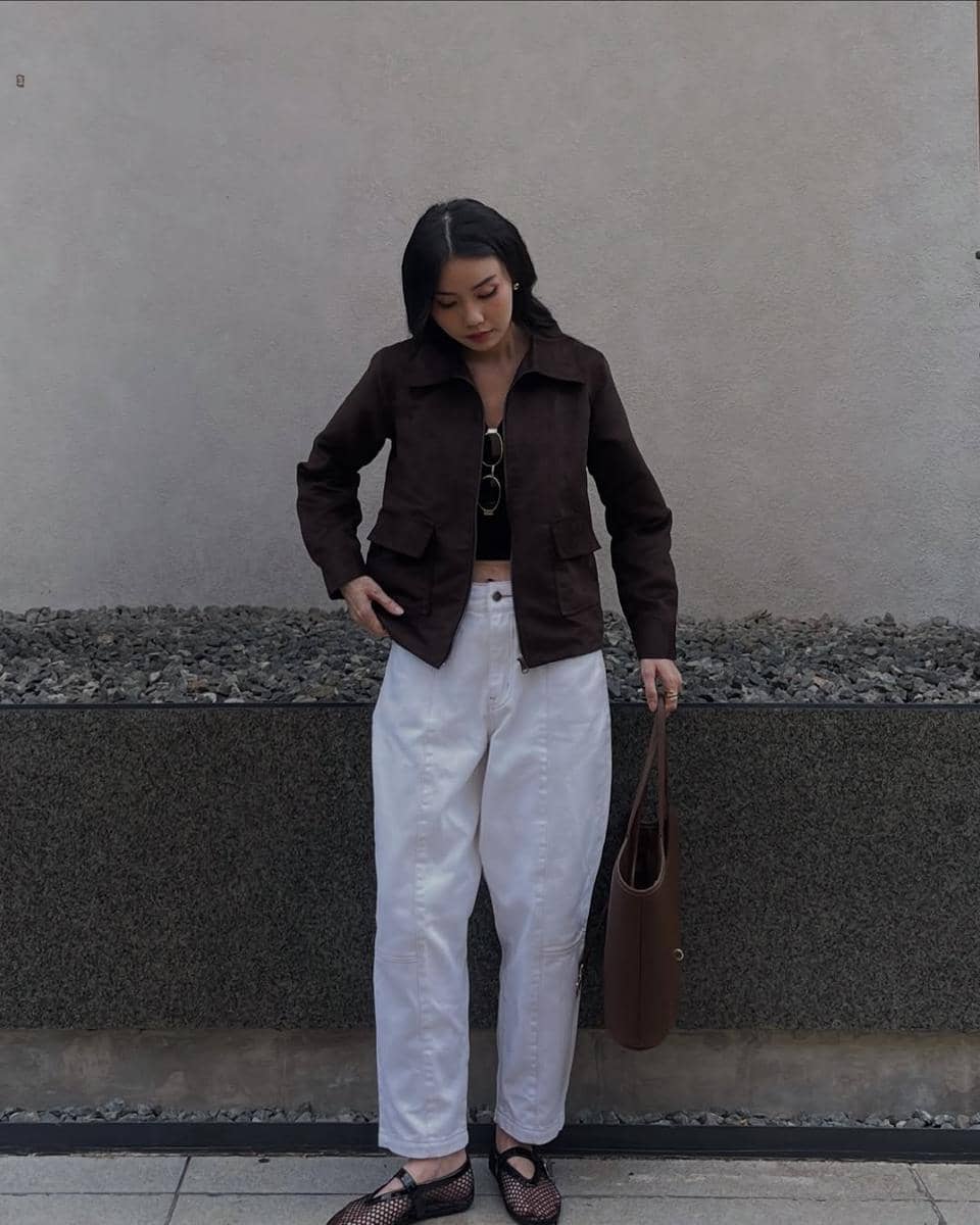 Seorang wanita berdiri di depan dinding abu-abu mengenakan jaket cokelat, celana putih longgar, dan membawa tas cokelat besar.