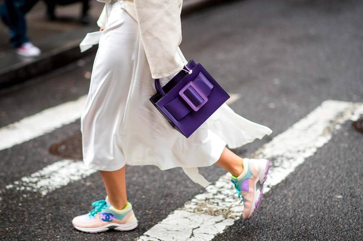 Seseorang berjalan di jalan dengan rok putih, sepatu kets warna pastel, dan tas tangan ungu dengan gesper besar.