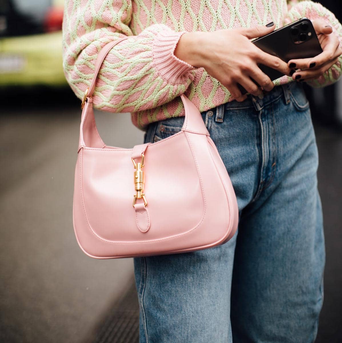Tas bahu kulit berwarna pink dengan detail gesper emas dipadukan dengan sweater rajut pastel dan celana jeans biru.