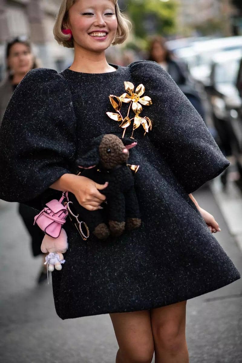 Seorang wanita tersenyum mengenakan gaun hitam berlengan besar dengan hiasan bunga emas dan membawa boneka anjing hitam di jalan kota.