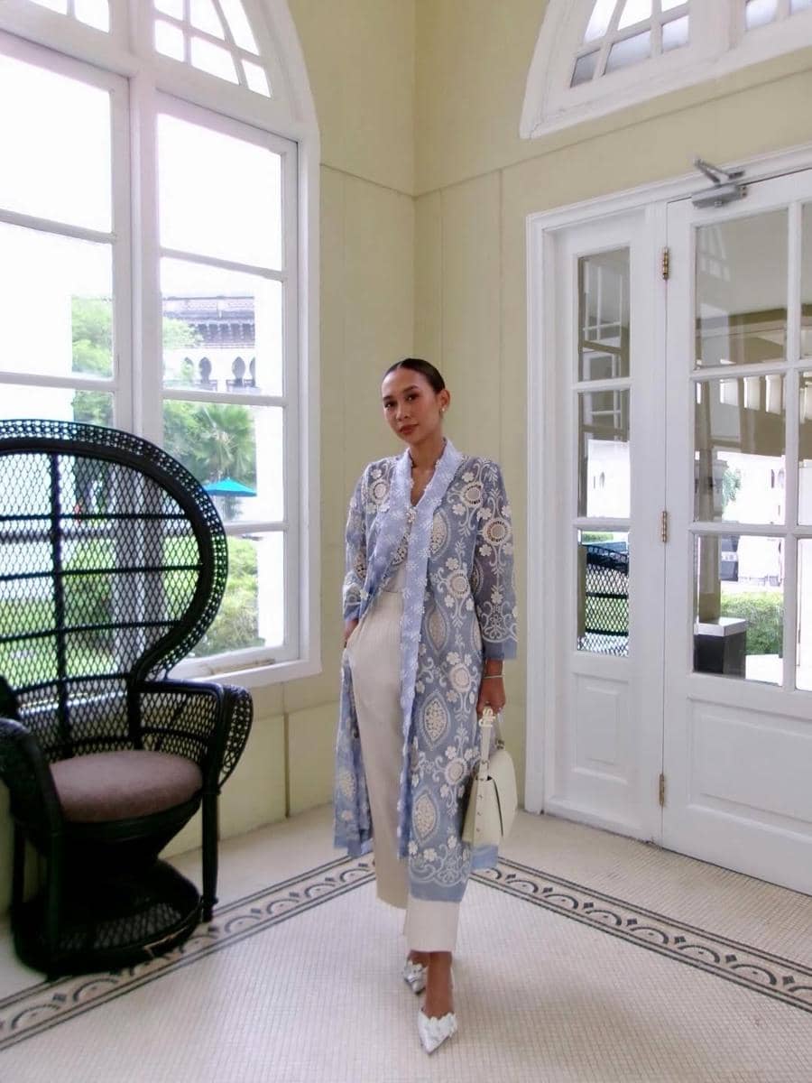 Seorang wanita berpose di dalam ruangan bergaya klasik dengan jendela besar, mengenakan outer biru bermotif dan celana krem.