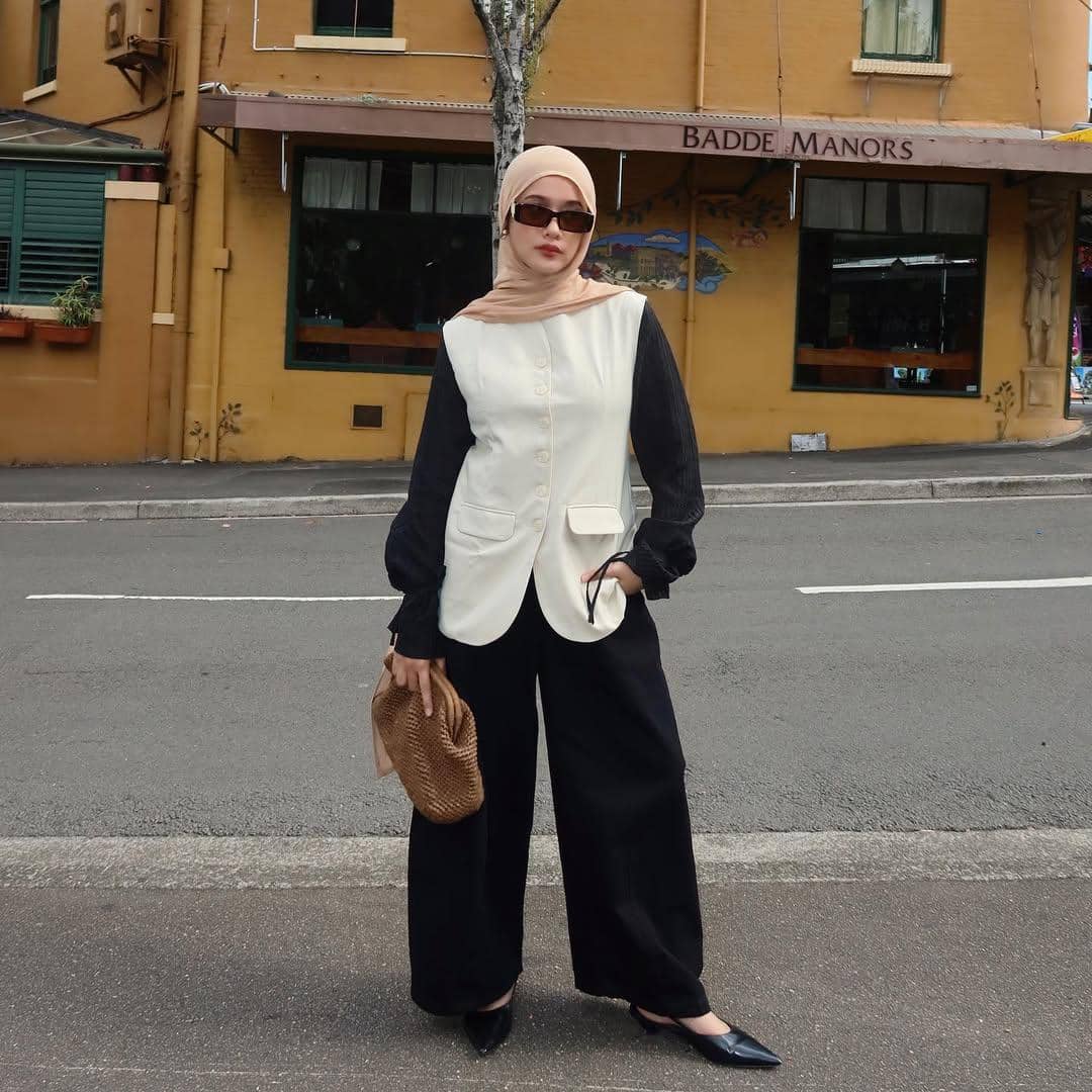 Seorang wanita berhijab mengenakan kacamata hitam, atasan krem dengan lengan hitam, celana panjang hitam, dan membawa tas cokelat di tepi jalan kota.
