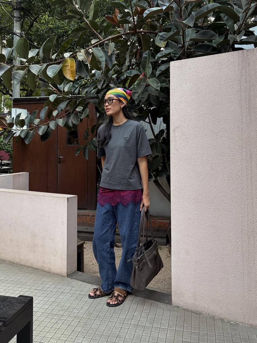 Seorang perempuan mengenakan kaus abu-abu, celana jeans longgar, dan bandana warna-warni sambil membawa tas di area luar ruangan.