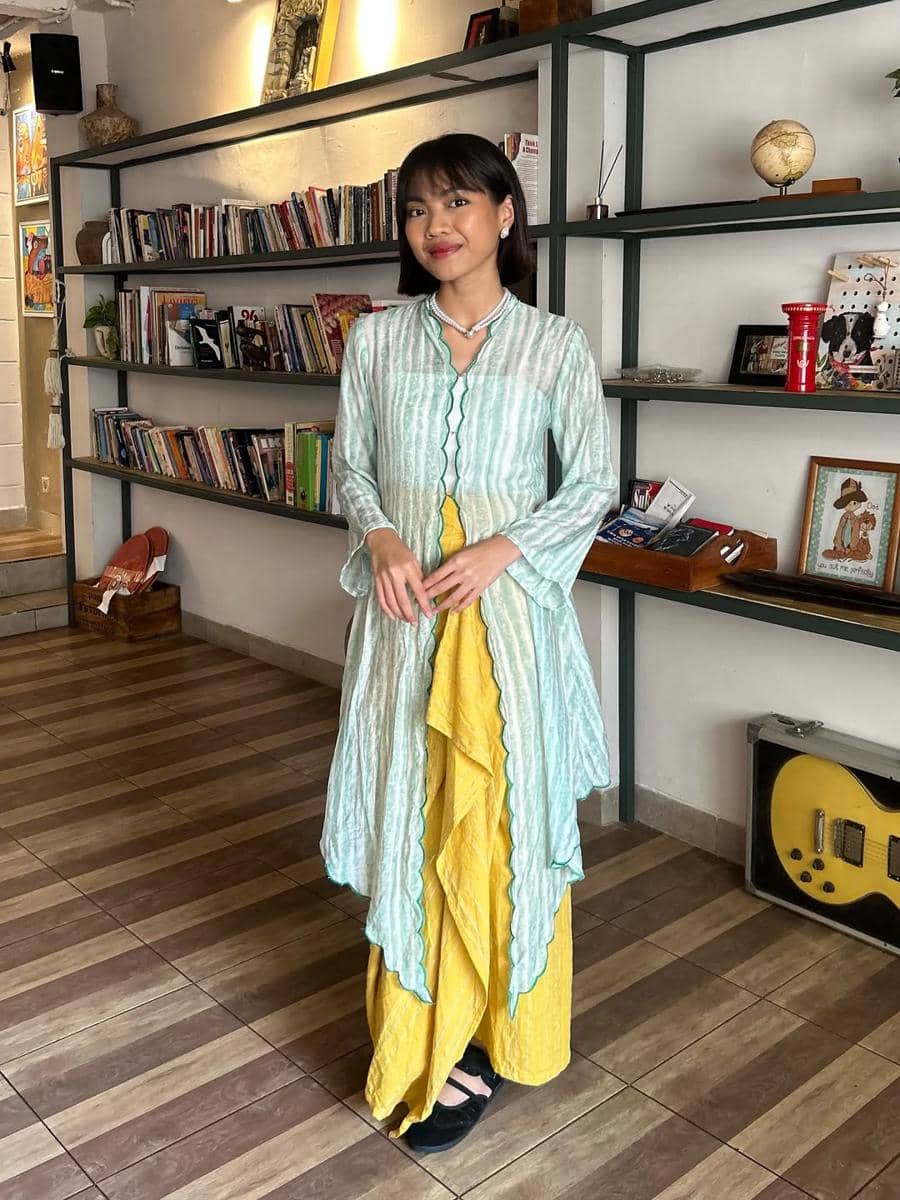 Seorang wanita mengenakan kebaya modern berwarna hijau muda dengan bawahan kuning berdiri di ruangan bergaya vintage dengan rak buku di belakangnya.