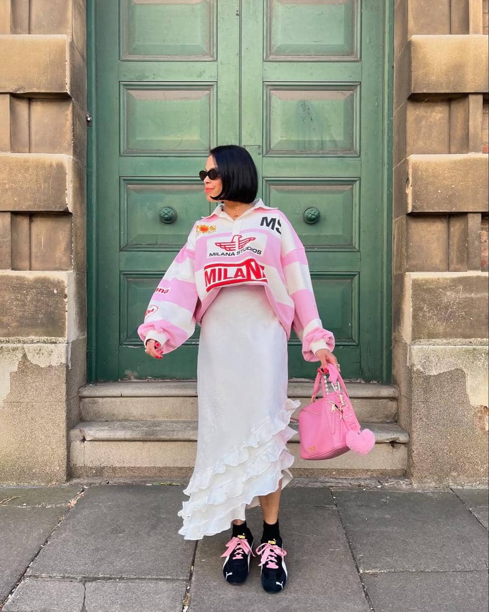 Seorang wanita berambut bob mengenakan sweater pink putih, rok putih berenda, dan sepatu hitam pink sambil memegang tas pink di depan pintu hijau besar.