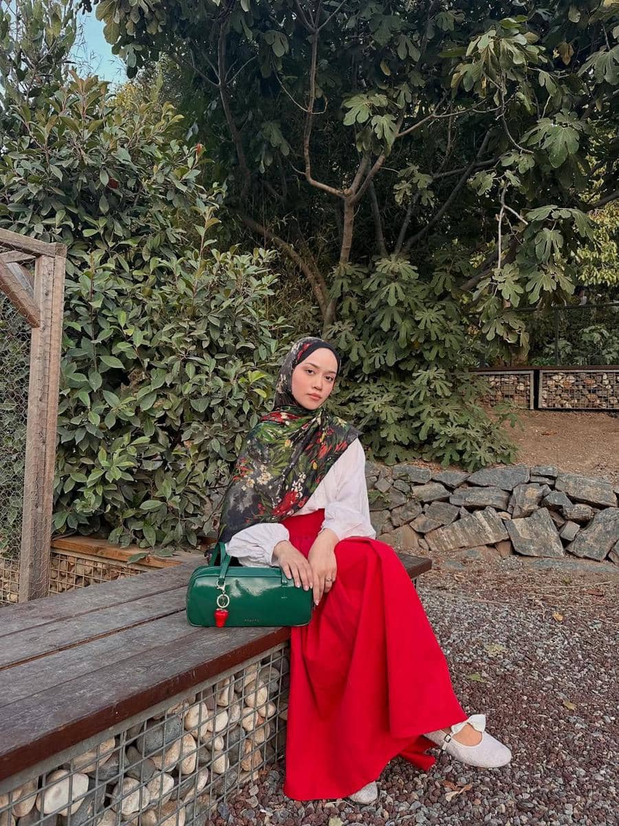 Seorang wanita berhijab duduk di bangku kayu di taman dengan latar pepohonan hijau, mengenakan rok merah dan membawa tas hijau.