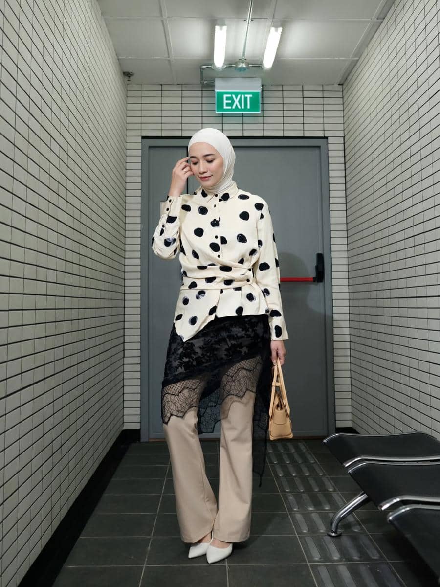 Seorang wanita berhijab mengenakan atasan motif polkadot krem dengan detail renda hitam, celana krem, dan tas kecil di lorong berubin putih.