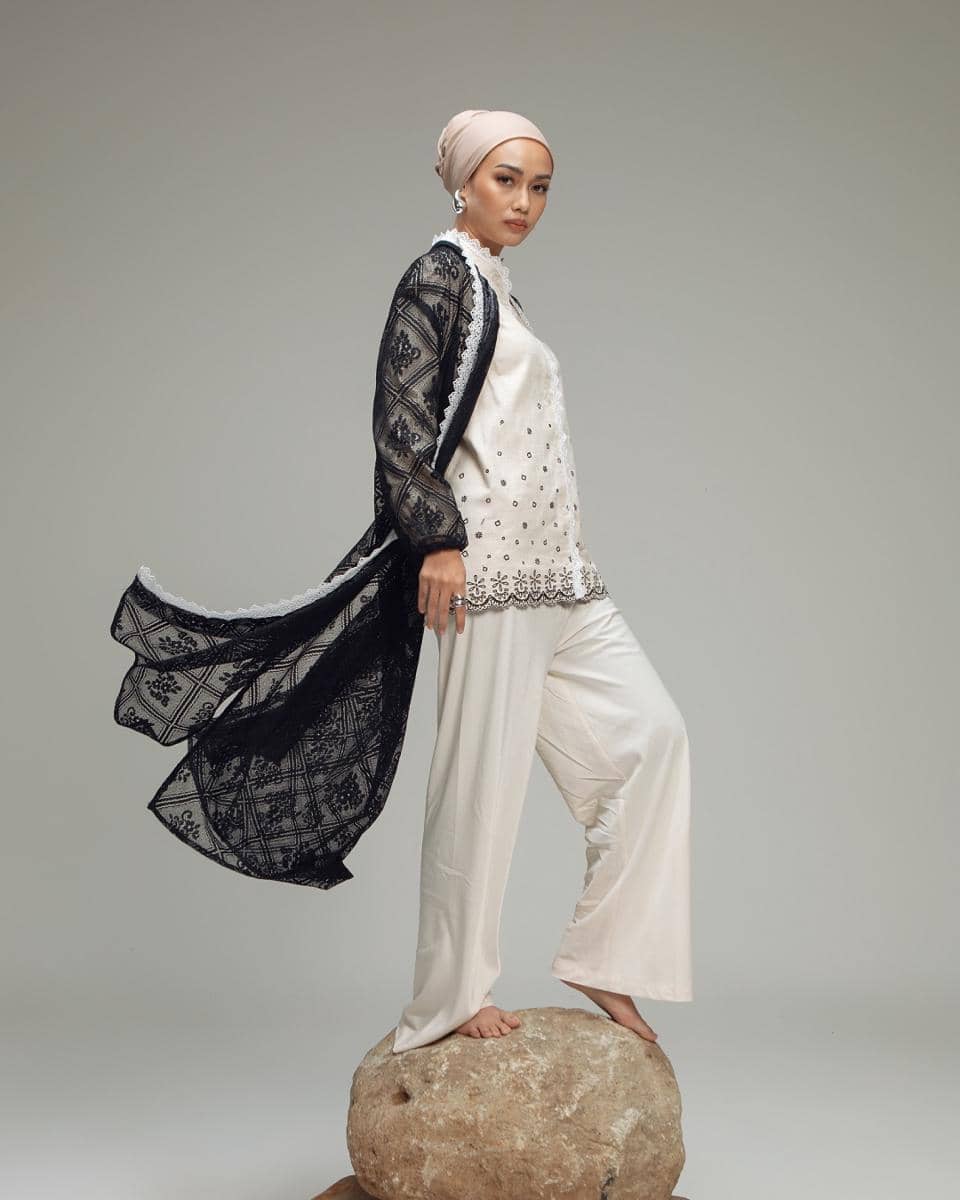 Perempuan berhijab mengenakan atasan putih berhias bordir dan outer hitam transparan berdiri di atas batu besar dengan latar abu-abu polos.